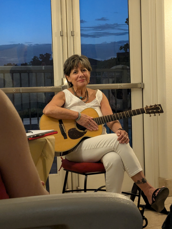 Photo of a seated woman holding a guitar in front of glass doors opening onto a porch at sunset